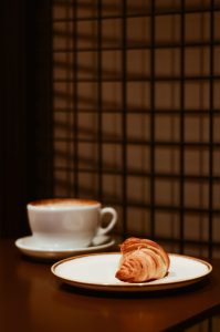 White coffee cup on a table with latte inside, and a white plate on the table with a croissant on it.