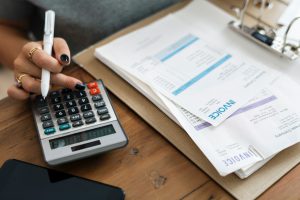 womans hand tapping a number on a calculator with invoices next to the calculator on a table.