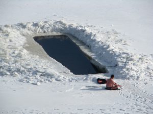 Caucasian man sitting in snow in front of a large rectangular opening of water dug out the snow. Cryotherapy.