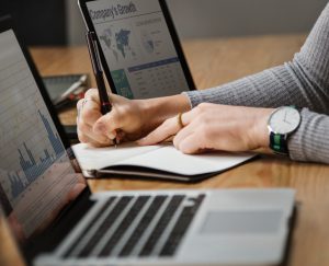 person writing in notebook between two laptops