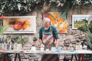 older caucasian man painting on a desk with paintings hung up behind him.
