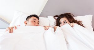 Asian man and womans faces peeking through a white comforter in bed.