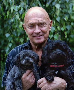 Older caucasian man smiling while holding up two black dogs.