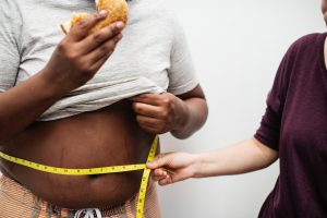 African American man lifting his white shirt reveiling his stomach with a caucasian woman measuring it with yellow tape.