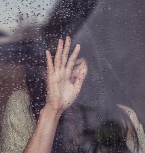 Caucasian woman crying with hand on glass that is wet from rain.