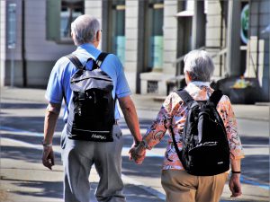 The back of an older man and woman with black backpacks on holding hands while walking.