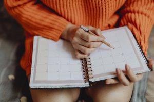 Caucasian woman's torso sitting down in an orange sweater with a planner on her lap writing in it.
