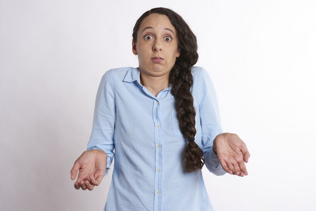 Caucasian woman with a side braid and light blue collared shirt on shrugging with her hands mid waist.