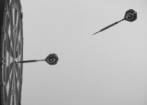 black and white photo of a dartboard with one dart in the center and another being thrown towards the center.