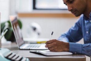 young man in blue shirt writing down in a notebook with a laptop open in the background.