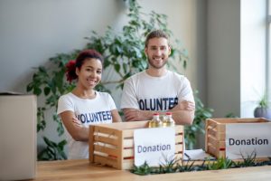 a man and woman with white shirts that say volunteer on them with a wodden box on a table that has a white sign with donation on it