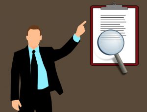 cartoon of a man in a suit pointing at a clipboard with magnifying glass over a certain part. 