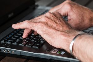 older caucasian hands typing on a laptop keyboard with a silver bracelet on one wrist.