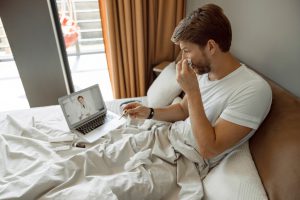 caucasian man in bed with a tissue in one hand and thermomter in the other with a laptop on the bed and a doctor on the screen.