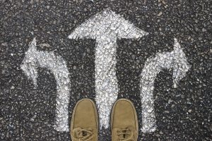 A pair of shoes stanfing in front of three white arrows on asphalt pointing in different directions.