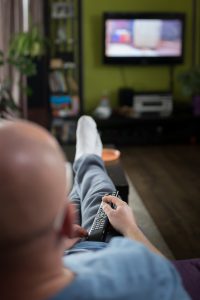 caucasian man laying on a couch with a remote in his hand with a blurred tv in the background.