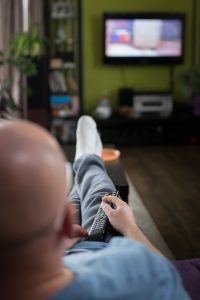 caucasian man laying on a couch with a remote in his hand with a blurred tv in the background.