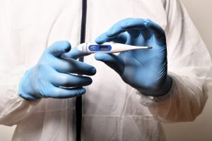 person in white coat with blue gloves on holding a thermometer in one hand and pointing at the audience with the other.