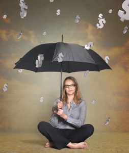 caucasian woman in business attire sitting under a black open umbrella with dollar signs coming down like rain.