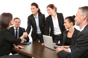 women and men in business suits sitting at a table, one woman standing up shaking the hand of a woman sitting down.