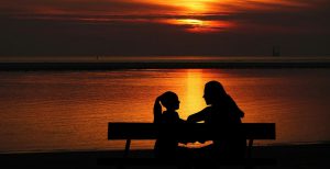 silhouette of a woman and an adolescent girl sitting on a bench facing each other with a sunset in the background.