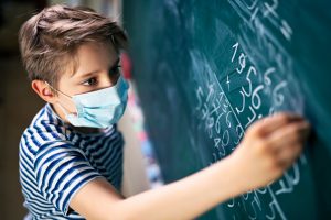 Caucasian boy with a mask on writing on the blackboard. 