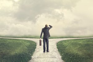 man in a business suit staring at a crossroad with one hand holding a suitcase and the other on his head.