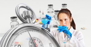 caucasian woman in a lab coat with gloves on mixing liquids with a stopwatch in front and vaccine bottles in the background.
