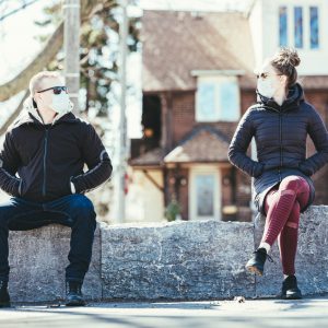 caucasian man and woman sitting 6 ft apart on a wall with masks on and hands in their pockets looking at each other.