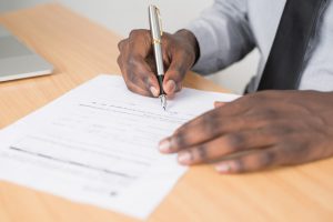 afircan american hands with one hand on a table and the other holding a gray pen signing the paper on a table.