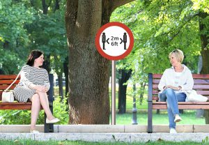 two caucasian women sitting on separate benches 6 ft apart with a sign in the middle that says 6 ft
