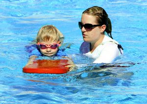 caucasian woman with glasses on and a whistle around her neck holding a water surfer with caucasian kid holding it. 
