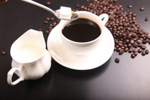 black coffee in a white up and saucer with a sugar cube in a spoon over the cup, and coffee beans on the table