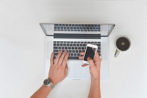 caucasian hands with one hand on laptop keyboard and the other hand holding a cell phone