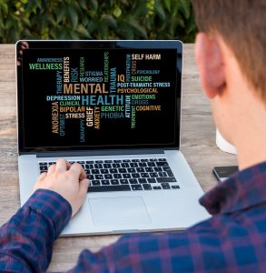 caucasian man sitting in front of a laptop with mental health related words on the screen.