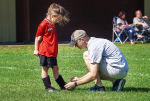 caucasian dad kneeling down to tie his daughters cleat., who is in soccer uniform.