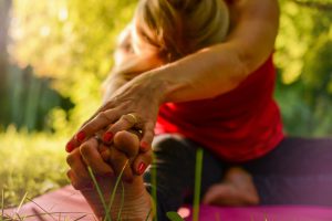 caucasian woman sitting on a yoga mat outside stretching and grabbing her feet.
