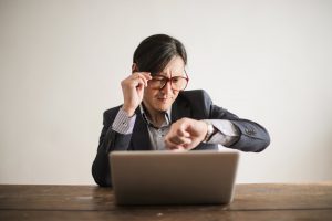 asian man looking at his watch while holding his glasses and sitting in front of a laptop