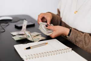 womans hands counting money at a table with a pen and notebook in front of her.