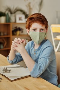 caucasian child with red hair sitting at a desk with a mask on