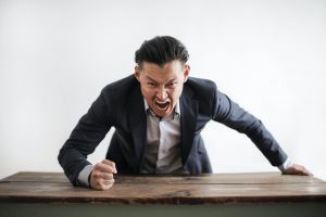 asian man in a suit standing up and pounding his fist on a table in rage.