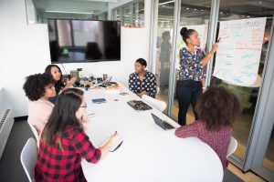 employees sitting around with one woman standing in front of a whiteboard pointing at it