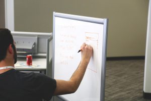 caucasian man sitting down writing on a white board.