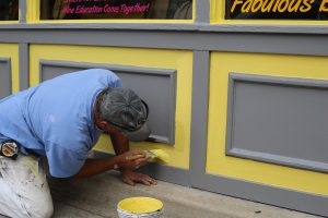 man kneeling down painting the outside of a building yellow and gray