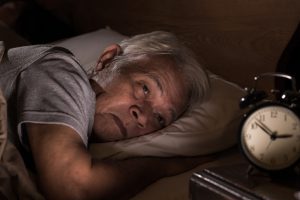 older asian man laying in bed staring at the clock on the table next to him.