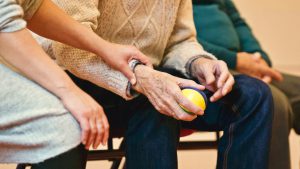 older mans lower half of body sitting down holding a ball in his hand with a person holding his arm in support