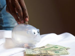 african american hand putting a coin into a clear piggy bank with money bills next to the piggy bank