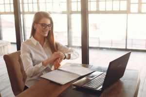 caucasian woman sitting in front of a laptop with a notebook in front of it and shes looking at her watch 