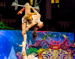 woman in a gold outfit hanging on a trapeze with one leg