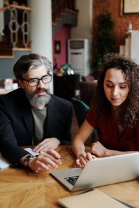 man in a suit pointing at a laptop screen with a woman next to him lookin at it